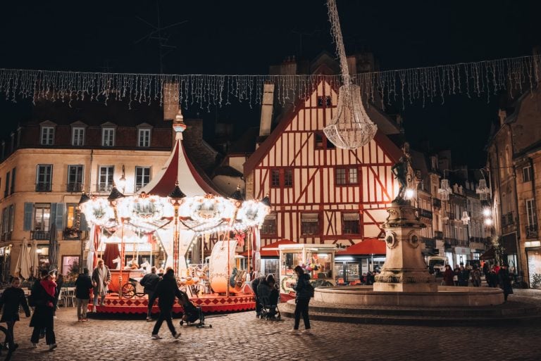 This is Place François Rude at night in Dijon. There is the infamous red half-timbered house and in front of it there is a fountain and a merry go round. People are walking around.