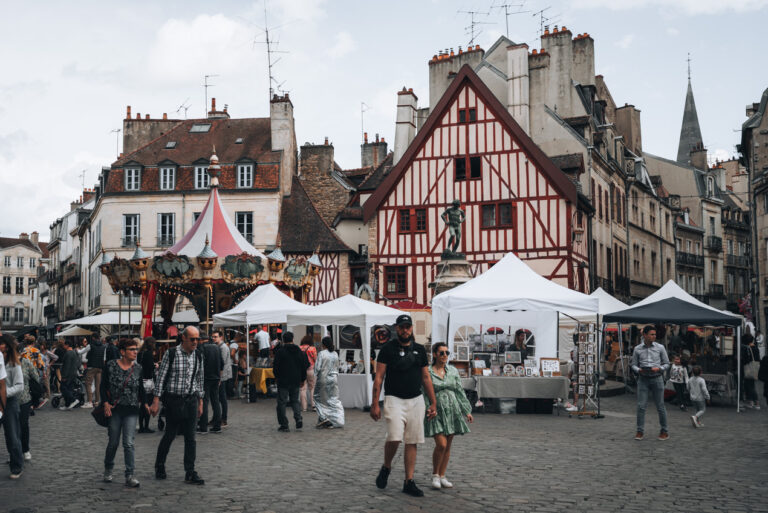 Outdoor market at Place François Rude in Dijon, with people browsing stalls beneath white tents, a red and white half-timbered house, carousel, and historic stone buildings in the background.