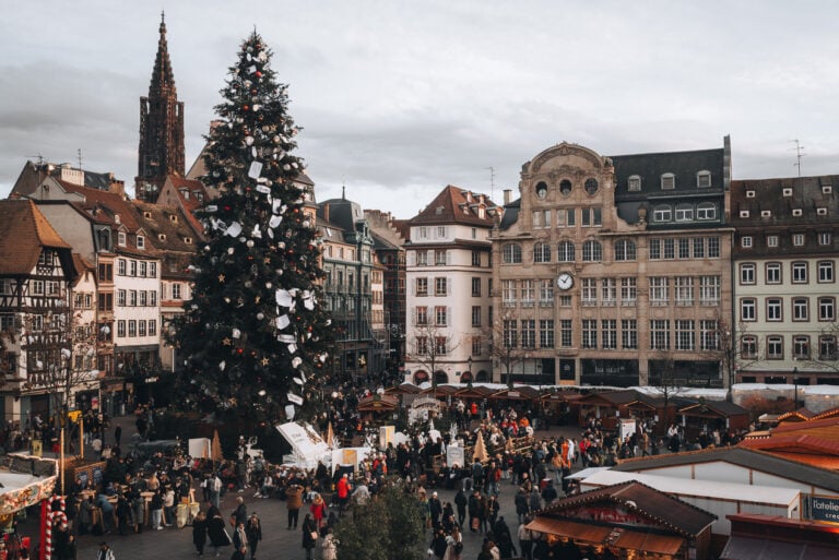 This is Place Kléber at the Strasbourg Christmas Market. There is a giant Christmas tree surrounded by wooden chalets. People are walking around and shopping.