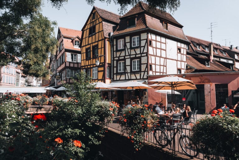 Sunlit summer view of Place de l’Ancienne Douane in Colmar with a central fountain surrounded by colorful half timbered buildings and outdoor café seating. This lively square is a key highlight in Colmar.