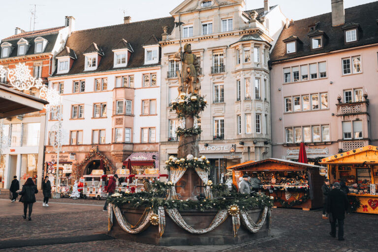 Place de la Réunion at the Christmas market in Mulhouse. There is the Fontaine du Hallebardier decorated in the Christmas cloth and surrounded by white stone buildings.