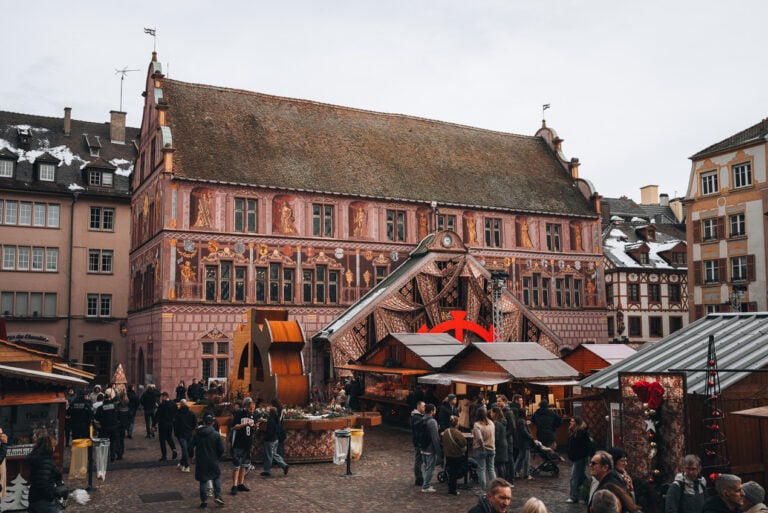 This is Place de la Réunion at the Mulhouse Christmas Market. There is the history museum decorated with the Christmas fabric and in front of it there are wooden chalets selling food and crafts.