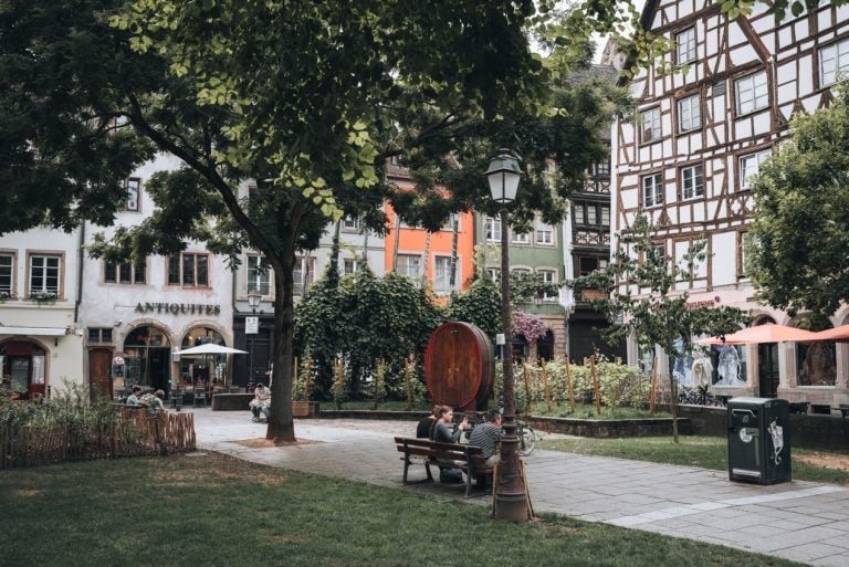 This is Place des Tripiers. It's a small square with benches and trees. There are buildings surrounding the square and people are sitting on the benches.