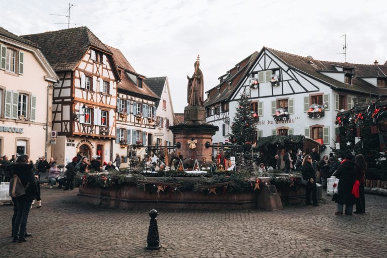 This is Place du Château Saint-Léon in Eguisheim during the Christmas market. There is the fountain decorated with wreaths and ornaments and half-timbered houses surrounding it.