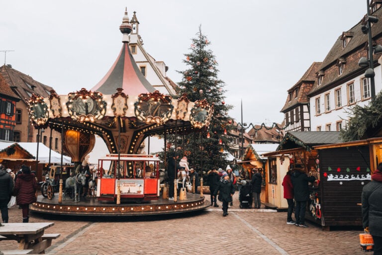 Festive scene at the Obernai Christmas market in Place du Marché with wooden stalls a large decorated Christmas tree and a carousel in the square. Visitors stroll through the market surrounded by holiday lights and decorations.