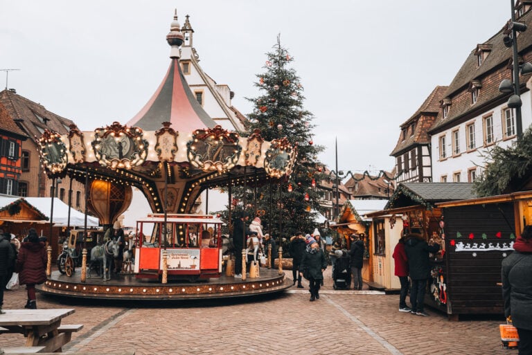 This is Place du Marché at the Obernai Christmas Market. There is a carousel, a Christmas tree, and several wooden chalets selling local products. 