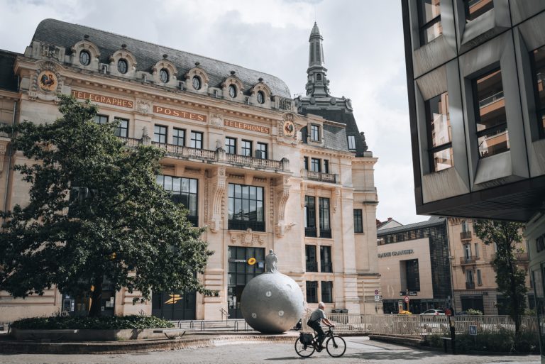 This is Poste Place Grangier in Dijon. It's a small square with the post office in the background. In front there is a tree, a circular statue, and a man riding a bike. 