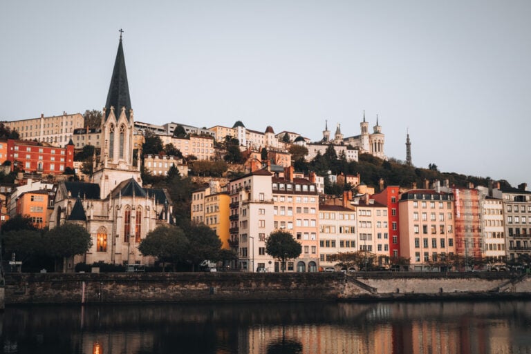 Quai Fulchiron in Vieux Lyon along the Saône River at sunrise with soft golden light reflecting off the water. The colorful Old Town buildings and St. George's Church offer a peaceful early morning view.