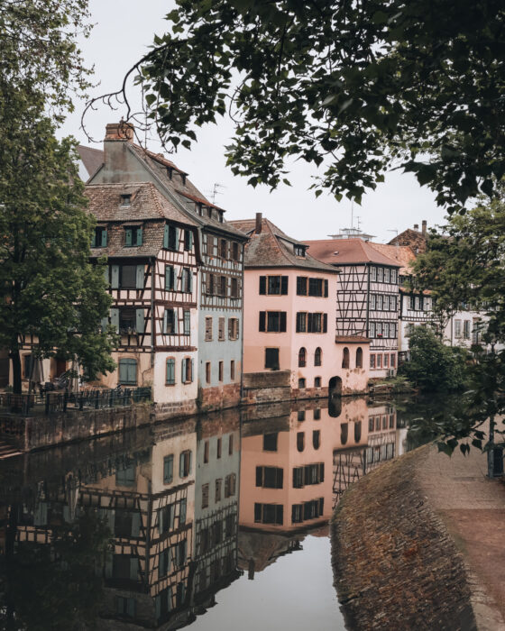 Sunlit canal walkway along Quai de la Bruche in Strasbourg lined with leafy green trees and historic buildings reflected in the calm water. The vibrant summer greenery and peaceful riverside path make it a scenic example an instagram spot during warmer months.