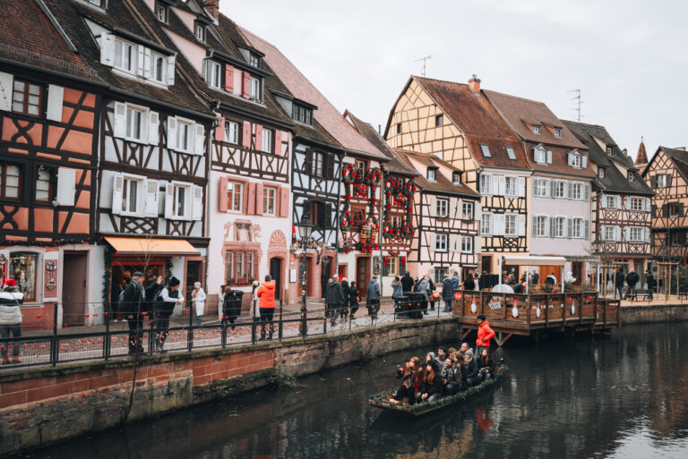 Pastel half-timbered houses along Quai de la Poissonnerie during Colmar's Christmas market. Each one is decorated with festive Christmas ornaments and lights.