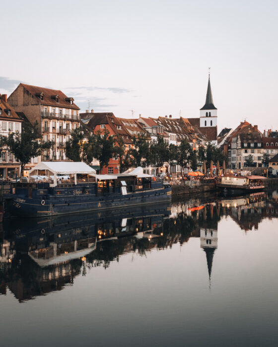 Warm sunset light reflects off the calm canal at Quai des Pêcheurs in Strasbourg, with boats and historic buildings lining the waterfront. The soft golden sky and tranquil water create a romantic evening scene.
