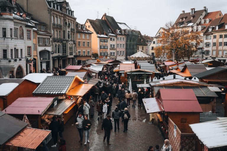 This is a rainy day during the Christmas market in Mulhouse. There are clouds in the sky and the ground is wet and people are walking around with jackets and hats.