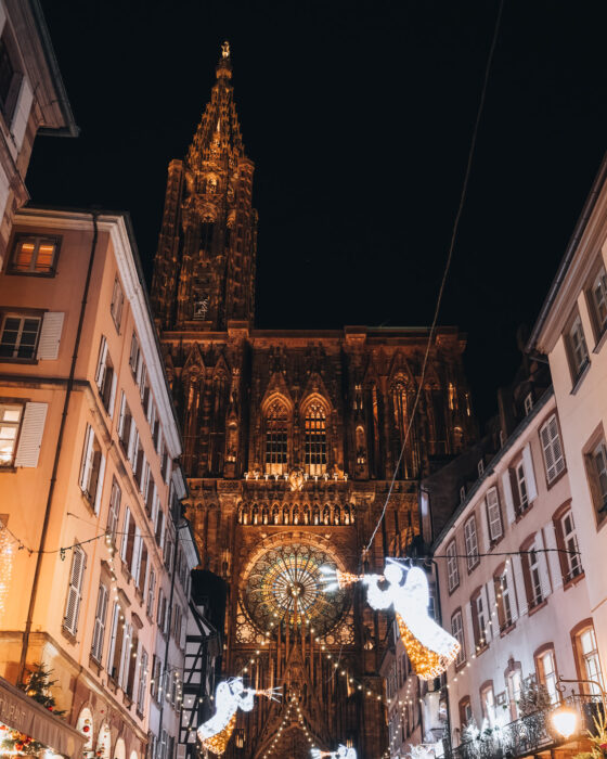 Strasbourg's Rue Mercière at night during the Christmas market, with stone buildings framing Strasbourg Cathedral and festive lights hanging from each side.