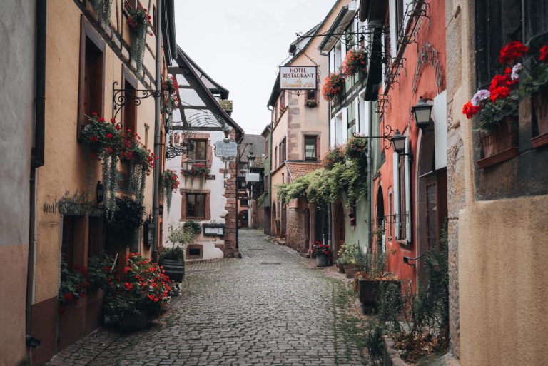 This is Rue du Cerf in Riquewihr. It's a cobblestone street and it's lined with colorful buildings. Each building has flowers and vines hanging down. It's one of the best day trips from Colmar.