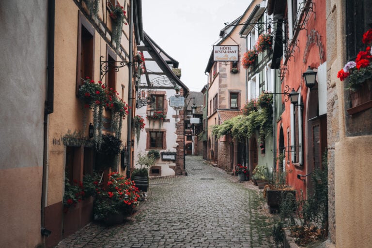 Rue du Cerf in summer in Riquewihr. The cobbled street is lined with colorful buildings and pots of overflowing flowers. It's a picture-perfect spot along the Alsace Wine Route.