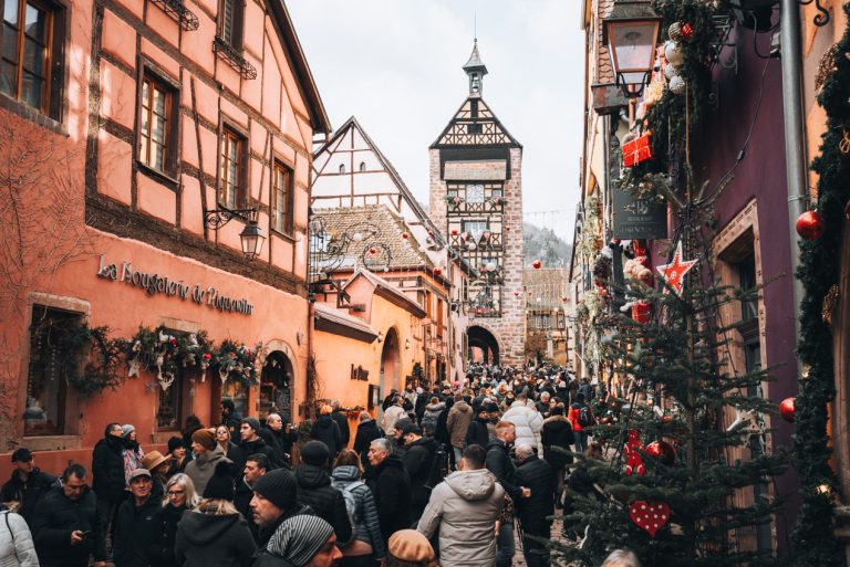This is Rue du Général de Gaulle at the Christmas market in Riquewihr. There is a mob of people walking along a street and the buildings are decorated with lights and ornaments. This is one reason not to visit Alsace.