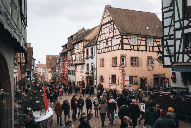 This is Rue du Général de Gaulle at the Riquewihr Christmas Market. There are colorful buildings decorated with lights and ornaments. The street is filled with people.