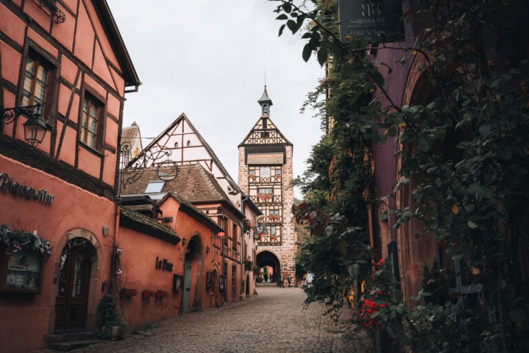 This is Rue du Général de Gaulle in Riquewihr. It's a cobbled street lined with colorful buildings and in the distance there is a watch tower.
