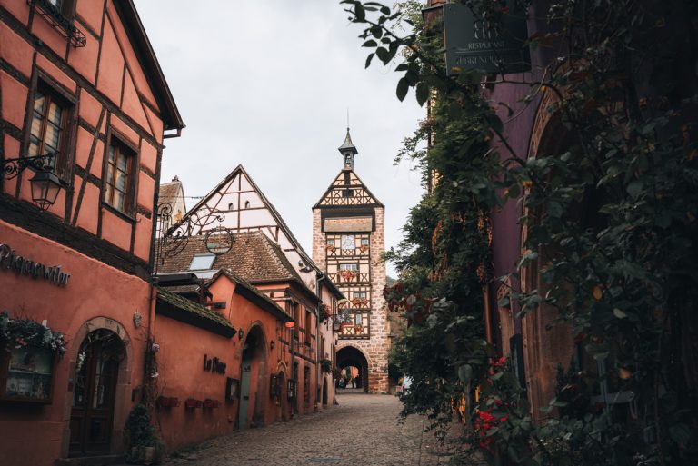 This is Rue du Général de Gaulle in Riquewihr. It's a cobblestone street lined with colorful building, each decorated with hanging vines. At the end of the street there is the watchtower with a clock.