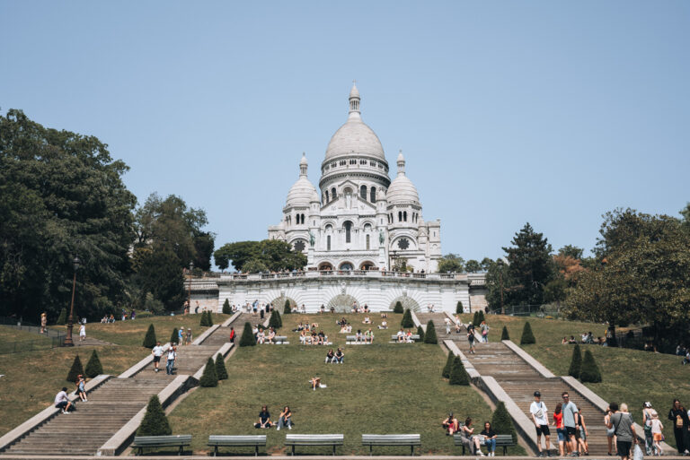 The Sacré-Cœur Basilica in Paris on a clear summer day, with people relaxing and socializing on the grassy hillside and stone steps leading up to the white domed church.