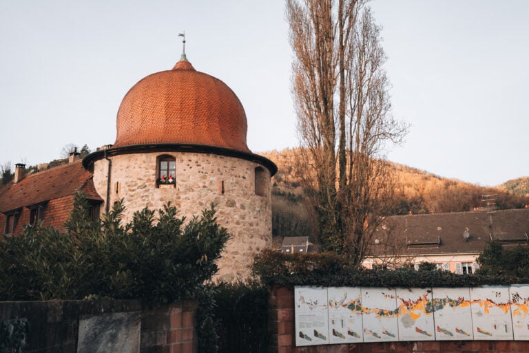 A tall stone tower surrounded by bushes marks the southern end of the Alsace Wine Route in Thann. The surrounding trees and bushes are brown which is what it looks like in winter.