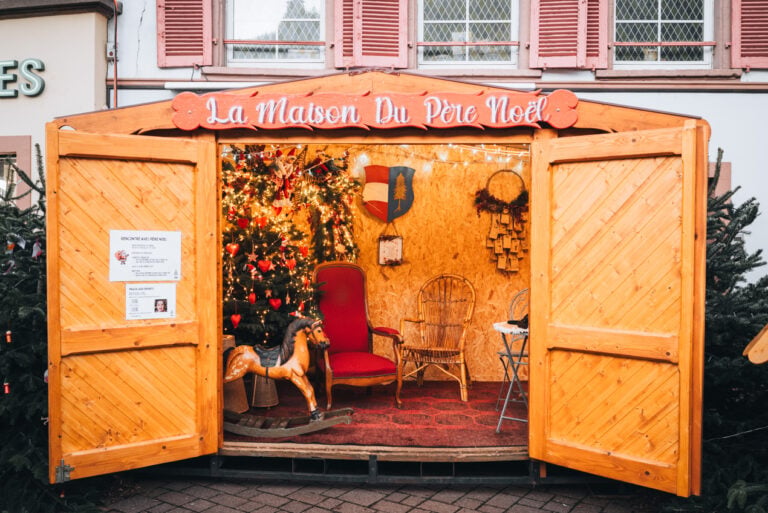 A wooden hut with The House of Santa written in French across the top at Place de Lattre de Tassigny in Thann. Inside there is a red chair, Christmas tree, and a wooden rocking chair.