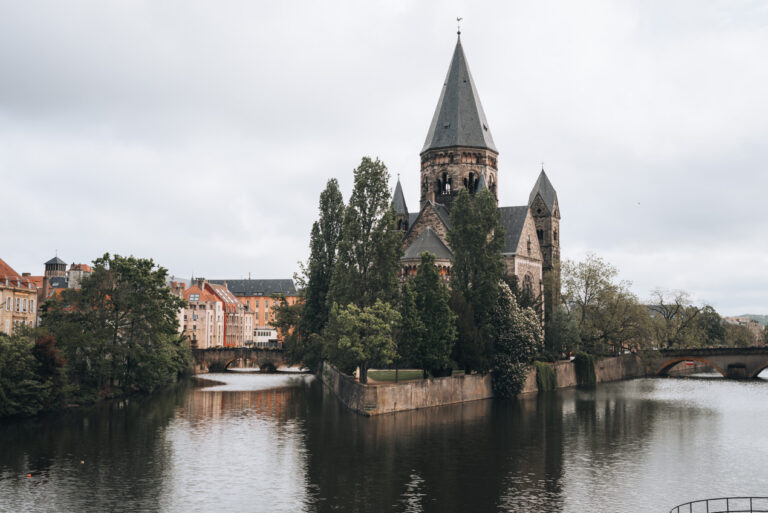 The Temple Neuf in Metz, a Romanesque Revival church with a tall central spire, stands on a tree-lined island surrounded by the Moselle River, with historic buildings and stone bridges in the background on a cloudy day.