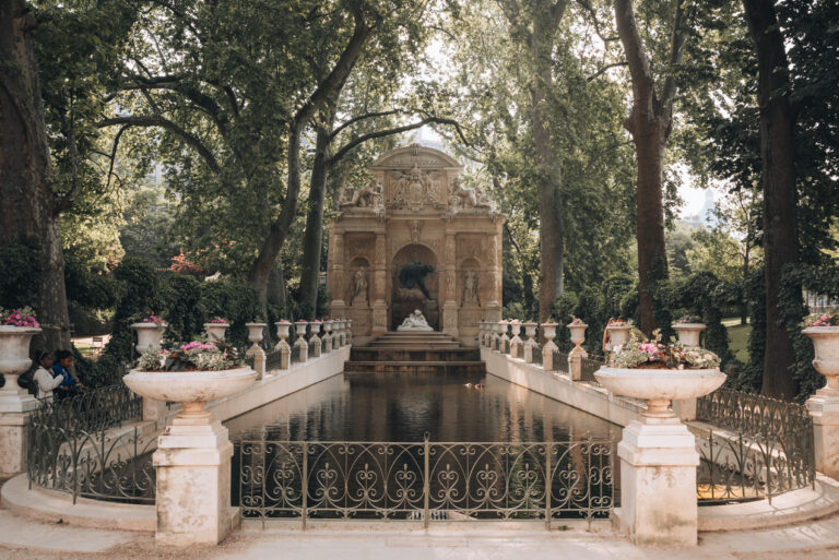 Medici Fountain surrounded by lush green trees and bouquets of rose flowers in the Jardin du Luxembourg. This is one of the my favorite things to do in Paris in summer.