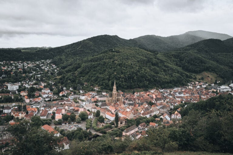 Bird's eye view of Thann's city center from the Croix du Rangen with the rooftop visible in the foreground and the tree covered rolling Vosges mountains in the distance. Hiking to this viewpoint is one of the best things to do in Thann.