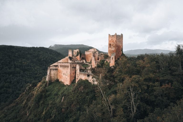 This is a view of Château de Saint-Ulrich and I took this photo from the terraces of Château du Giersberg in Ribeauvillé Alsace. The castle is made of red stone and it's surrounded by trees. This hike is one of my favorite things to do in Ribeauvillé.