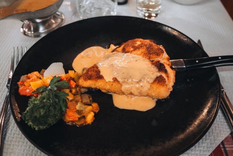 Plate of traditional Alsatian cordon bleu served with golden fried crust, melted cheese, and ham inside, alongside a side of potatoes. The hearty regional dish reflects the food culture along the wine route in Alsace.