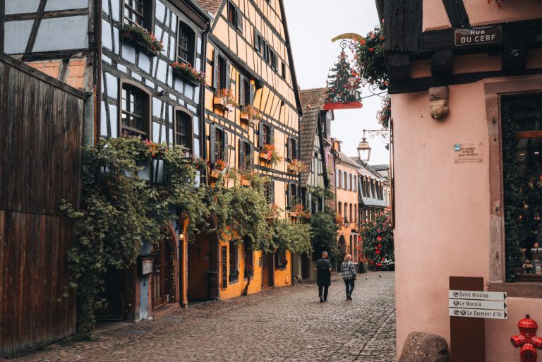 This is a row of traditional houses in Riquewihr. They are all different colors and are draped in hanging vines. There are two people walking down the cobble street.