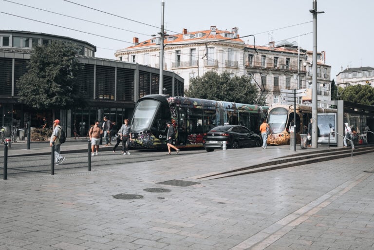 This is a photo of two trams parked at the Observatoire tram station in Montpellier. There are people walking raound and getting on and off each tram.