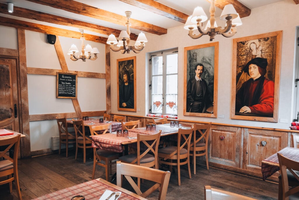 Cozy upstairs dining room at Le Fer Rouge in Colmar with wooden beams, warm lighting, and closely set tables ready for guests. The traditional Alsatian restaurant atmosphere reflects the traditional Alsatian restaurants.