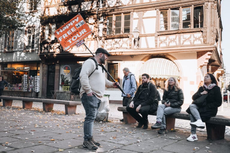 Vincent from Happy Strasbourg on the Original Free Tour talking about the history of Place Saint-Etienne. He is holding a sign advertising the tour and people are listing to him talk.