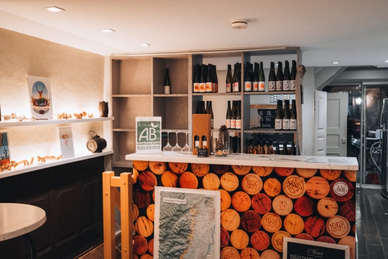 Wine Tasting Room at Domaine Martin Jund. There is a welcome counter with bottles of wine on shelves and tables and chairs.
