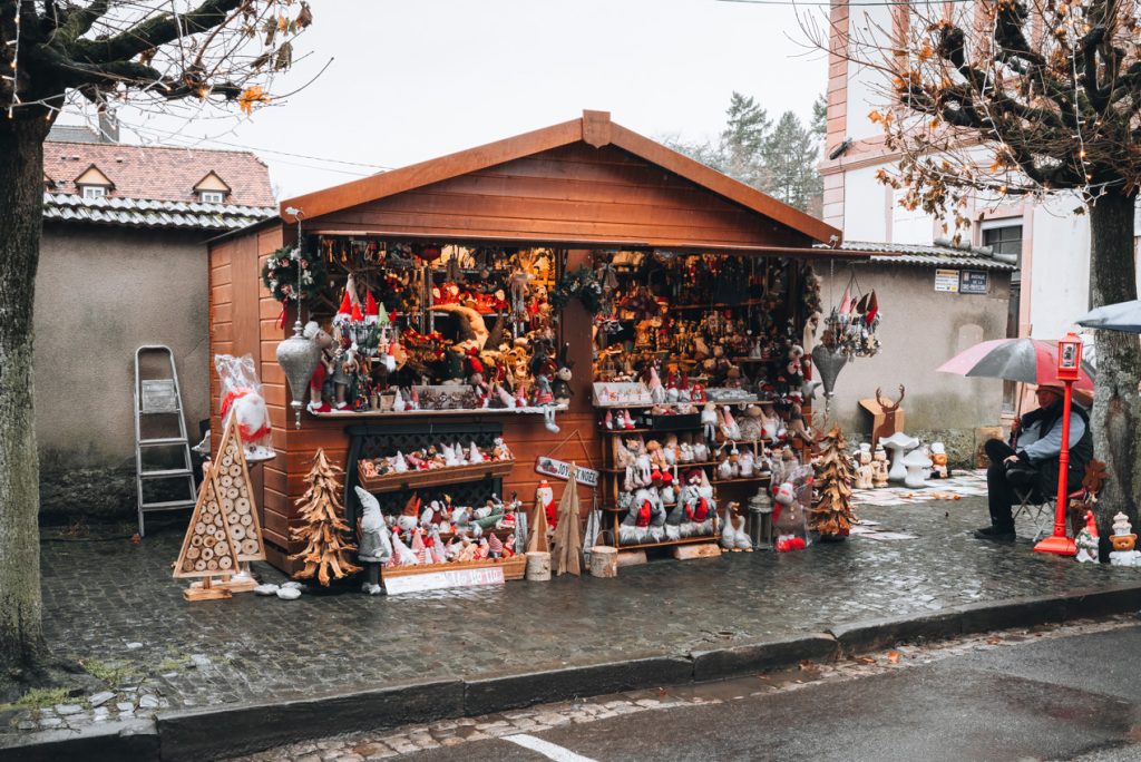 This is a wooden chalet at the Christmas market in Wissembourg. It is selling handmade crafts and the owner is sitting on the right hand side holding and umbrella.